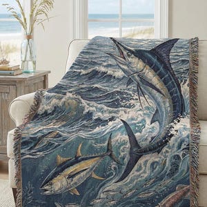 May include: A woven throw blanket featuring a marine scene with a marlin leaping from the ocean. The design includes other fish, waves, and a squid, in shades of blue, tan, and white. The blanket has a fringed edge and is draped over a cream-colored armchair.