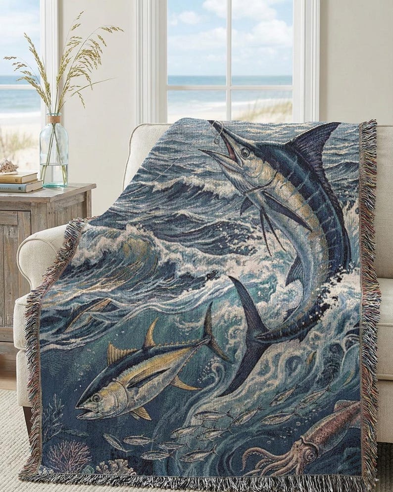 May include: A woven throw blanket featuring a marine scene with a marlin leaping from the ocean. The design includes other fish, waves, and a squid, in shades of blue, tan, and white. The blanket has a fringed edge and is draped over a cream-colored armchair.
