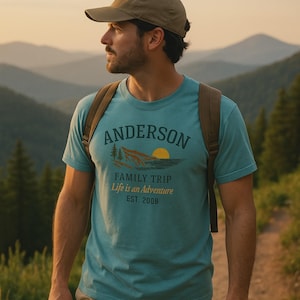 Op de afbeelding: Tealkleurig T-shirt met de naam "Anderson" boven een berg- en zongrafiek. Onder de afbeelding staat de tekst "Family Trip Life is an Adventure EST. 2008". De persoon draagt een bruine rugzak en een groene pet.