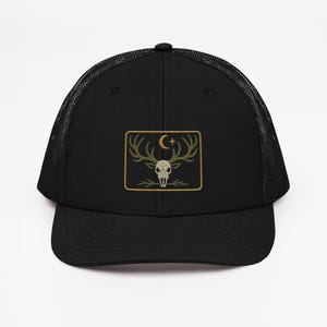 May include: Black trucker hat with a mesh back and a curved brim. The front features an embroidered patch with a deer skull, antlers, a crescent moon, and a star, all in shades of green and gold.