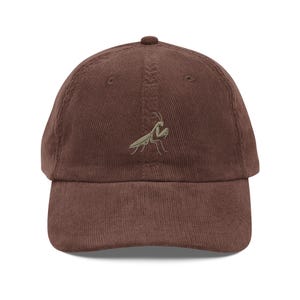 Puede incluir: Una gorra de béisbol de pana marrón con una mantis religiosa verde bordada en la parte delantera.