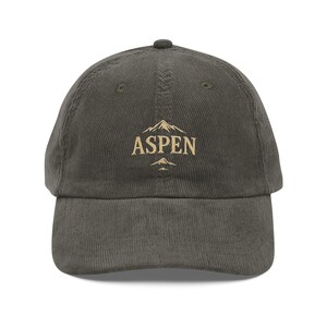 Könnte beinhalten: Olivgrüne Cord-Baseballkappe mit dem Wort "ASPEN" in goldener Schrift über einer Berg-Grafik. Die Kappe hat einen gebogenen Schirm und einen kleinen Knopf oben. Ein modisches Accessoire für Outdoor-Enthusiasten.