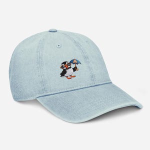 Puede incluir: Gorra de béisbol de mezclilla azul claro con un frailecillo de dibujos animados bordado que lleva gafas y sostiene un paraguas azul. El frailecillo tiene plumas negras y blancas, patas naranjas y un pico amarillo. La gorra tiene una visera curva.