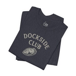 Peut inclure: Un t-shirt bleu foncé avec le texte "Dockside Club" en lettres blanches. Le t-shirt présente également un petit graphique blanc d'une coquille de palourde.