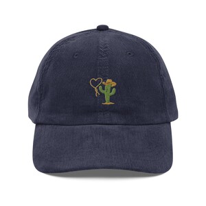 Puede incluir: Una gorra de béisbol de pana azul marino con un cactus bordado que lleva un sombrero de vaquero y sostiene un lazo en forma de corazón.
