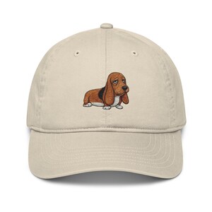 May include: A beige baseball cap with an embroidered cartoon basset hound with brown and white fur.
