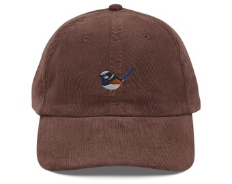 May include: A brown corduroy baseball cap featuring an embroidered bird design. The bird has blue, white, and orange details. The cap has a curved brim and an adjustable strap.