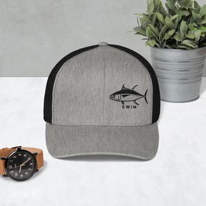 May include: A grey and black trucker hat with a black illustration of a fish and the word "SWIM". The hat has a curved peak and a mesh back. A watch with a brown leather strap is visible.