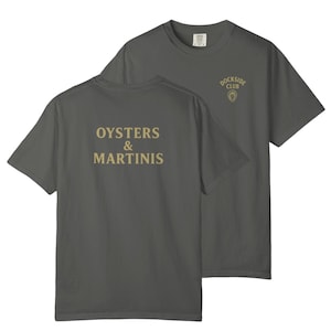 Peut inclure: Un t-shirt gris anthracite avec le texte "Oysters & Martinis" imprimé au dos en lettres dorées. Le devant du t-shirt arbore un petit logo doré qui dit "Dockside Club".