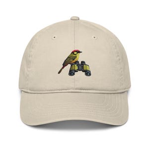 Op de afbeelding: Een beige baseballpet met een geborduurd beeld van een gele vogel die op een verrekijker zit.