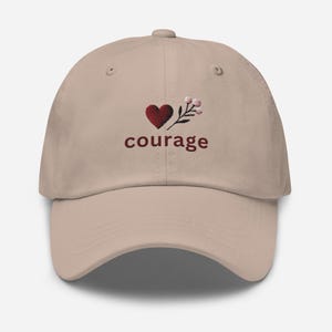 Puede incluir: Gorra de béisbol beige con visera curva. La gorra presenta un diseño bordado de un corazón rojo, una ramita de flores y la palabra "courage" en rojo. La gorra tiene un botón en la parte superior.