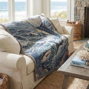 May include: A woven throw blanket featuring a marine life design, draped over a cream-colored sofa. The blanket showcases a detailed illustration of a marlin, tuna, and other sea creatures in shades of blue, green, and white. The sofa is in a coastal-themed living room.