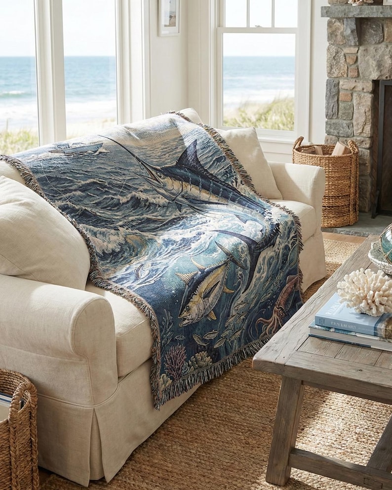 May include: A woven throw blanket featuring a marine life design, draped over a cream-colored sofa. The blanket showcases a detailed illustration of a marlin, tuna, and other sea creatures in shades of blue, green, and white. The sofa is in a coastal-themed living room.