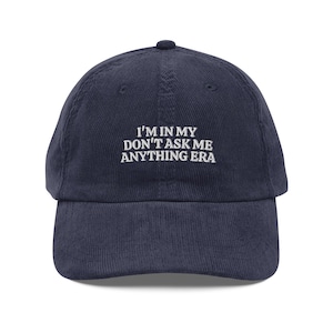 May include: Navy blue corduroy baseball cap with the embroidered text "I'M IN MY DON'T ASK ME ANYTHING ERA" in white. The cap has a curved peak and a rounded crown. A casual accessory.