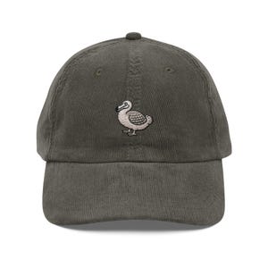Könnte beinhalten: Olivgrüne Cord-Baseballkappe mit einem kleinen, gestickten Dodo-Vogel-Motiv. Der Vogel ist in Weiß- und Beigetönen dargestellt. Die Kappe hat einen gebogenen Schirm und einen verstellbaren Riemen.