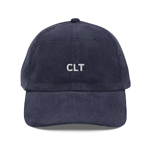 May include: A navy blue corduroy baseball cap featuring the white embroidered letters "CLT". The cap has a curved peak and a rounded crown. A classic style hat.