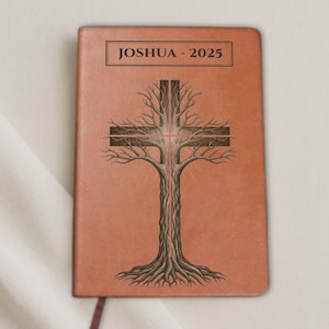 Puede incluir: Un diario de cuero marrón con el texto "JOSHUA - 2025" en la parte superior. La portada presenta una ilustración detallada de una cruz formada por un árbol, con raíces y ramas. Una cinta de marcador burdeos es visible.