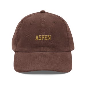 Op de afbeelding: Een bruine corduroy baseballpet met het woord "ASPEN" in goud geborduurd op de voorkant.