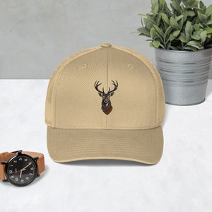May include: A tan baseball cap with a brown deer head embroidered on the front. The deer has antlers and a red ribbon around its neck.
