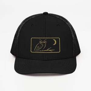 May include: Black trucker hat with a mesh back and a curved brim. The front panel features a rectangular patch with a gold embroidered owl perched on a branch, with a crescent moon in the background.