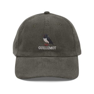 Op de afbeelding: Olijfgroene corduroy baseballpet met een geborduurd Guillemot vogelontwerp. De pet heeft een gebogen klep en een verstelbare band. De vogelillustratie is in zwart, wit en rood, met het woord "GUILLEMOT" eronder geborduurd.