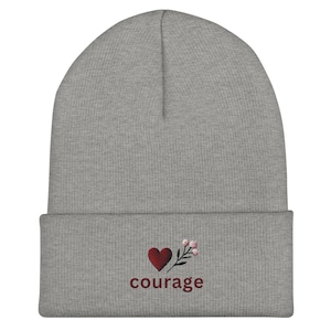 Puede incluir: Gorro gris con un corazón rojo y un pequeño tallo de flor bordados en la parte delantera. El texto "coraje" está bordado debajo del corazón.