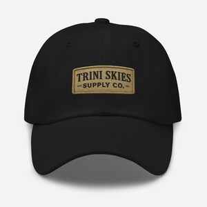 Può includere: Cappellino da baseball nero con una toppa marrone che dice "TRINI SKIES - SUPPLY CO.-"
