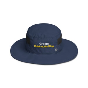 May include: A navy blue sun hat with a black mesh panel and the text "Groom Catch of the Day" embroidered in white and yellow.
