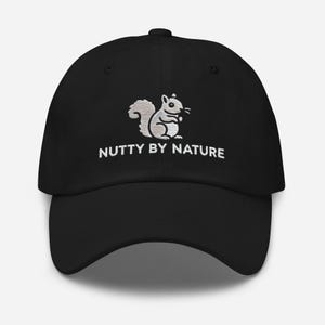 Op de afbeelding: Zwarte baseballpet met een wit geborduurd eekhoorn en de tekst "Nutty by Nature".