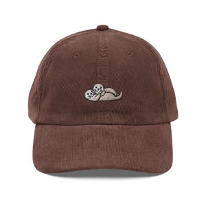 May include: A brown corduroy baseball cap with an embroidered image of two otters cuddling.