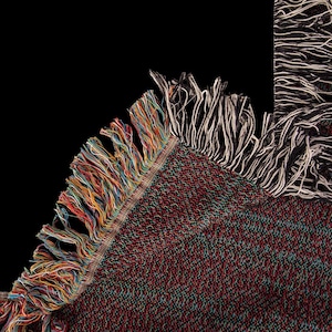 May include: Close-up of a woven blanket with a dark red, green, and black speckled pattern. The blanket has a colorful fringe of red, orange, yellow, blue, and green threads along one edge. A section of the blanket with white and black fringe is visible.