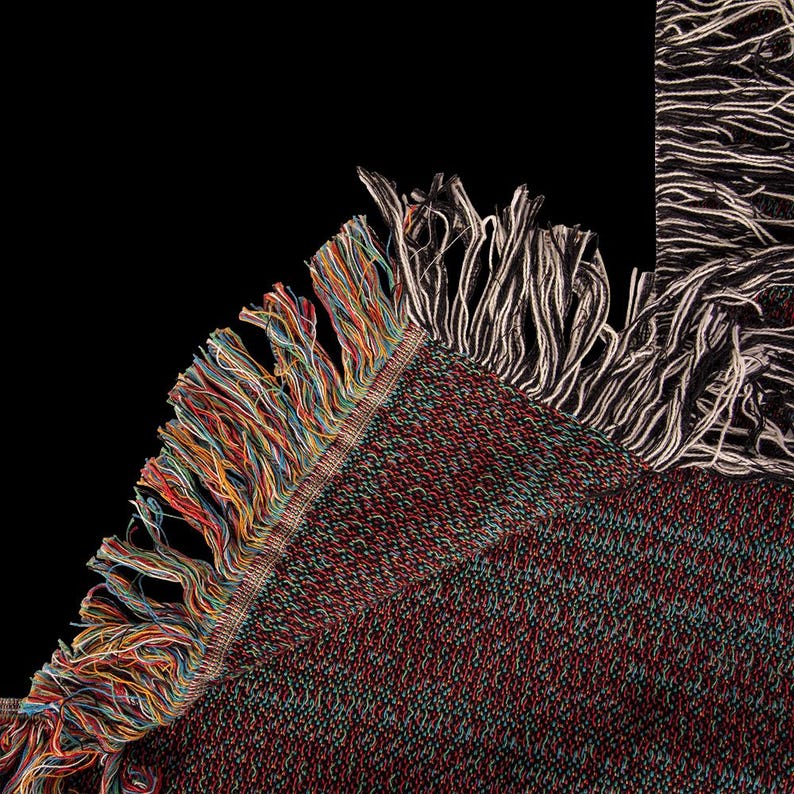 May include: Close-up of a woven blanket with a dark red, green, and black speckled pattern. The blanket has a colorful fringe of red, orange, yellow, blue, and green threads along one edge. A section of the blanket with white and black fringe is visible.