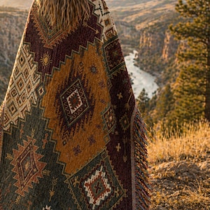 May include: A colorful woven blanket with a geometric pattern in shades of green, gold, and burgundy. The blanket has a fringed edge and is draped over a person, with a scenic background of a river and trees.