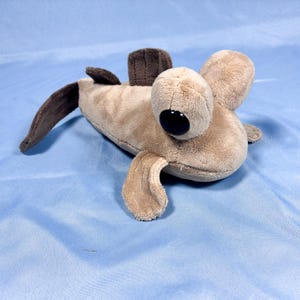 May include: A plush toy shaped like a mudskipper fish, featuring a light tan body with dark brown fins and a tail. The stuffed animal has large, prominent eyes with black pupils and a soft, velvety texture throughout its entire exterior surface.