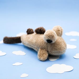 May include: A plush toy shaped like a creature with a tan, fuzzy body, dark brown fins, and a dark brown tail. The toy features two large, round, tan plush eyes with small black pupils, positioned on the top of its head, and a soft, textured surface.