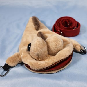 Puede incluir: Un bolso de peluche beige con forma de criatura, grandes ojos negros, un detalle de columna vertebral marrón oscuro y una abertura de boca con cremallera roja. Incluye anillas metálicas en los laterales y una correa roja ajustable.