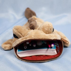 May include: A tan plush pencil case shaped like a creature with a wide zippered mouth. The interior is lined with red fabric and contains a spiral-bound notebook and several colorful pens. The exterior features soft fur, dark eyes, and a tail.