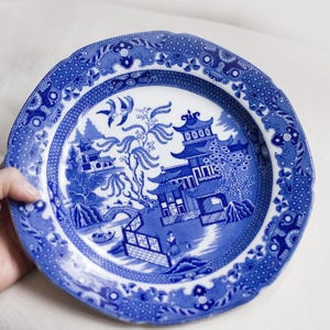 Puede incluir: Plato de cerámica azul y blanco con diseño Willow. El plato presenta una escena detallada de paisaje con pagodas, puentes y figuras. El borde tiene un borde festoneado y un borde decorativo. Una pieza clásica de vajilla.