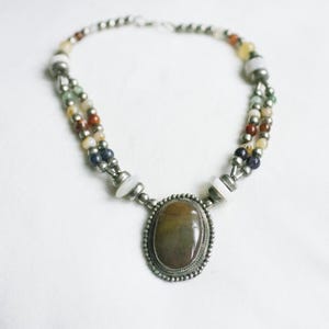 Puede incluir: Un collar de múltiples hilos con un gran colgante ovalado. El colgante es una piedra marrón y beige engastada en un metal plateado. Los hilos están hechos de pequeñas cuentas de colores en varios colores, incluyendo blanco, azul y rojo.