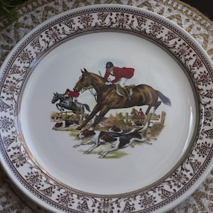 Puede incluir: Un plato de cerámica blanca con un borde floral marrón. El centro presenta una escena de caza con jinetes a caballo saltando una valla, acompañados de perros. Los jinetes visten chaquetas rojas.