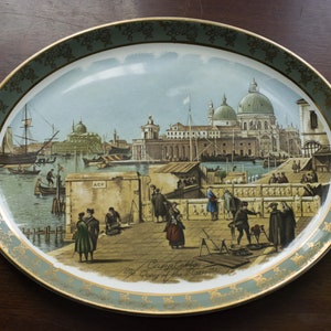 May include: A decorative oval ceramic plate with a green and gold border. The plate features a painted scene of a cityscape with buildings, boats, and people. The text "Canaleto The City of the Canals" is printed on the plate.