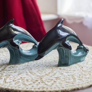 Puede incluir: Dos figuritas de delfines de cerámica, una negra y otra azul, saltando fuera del agua. Los delfines están sobre una base de ola azul y verde.