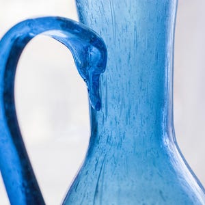 May include: A close-up of a blue glass jug with a curved handle. The jug has a textured surface and a wide opening. The handle is a darker shade of blue than the body of the jug. The background is a soft, blurred white.
