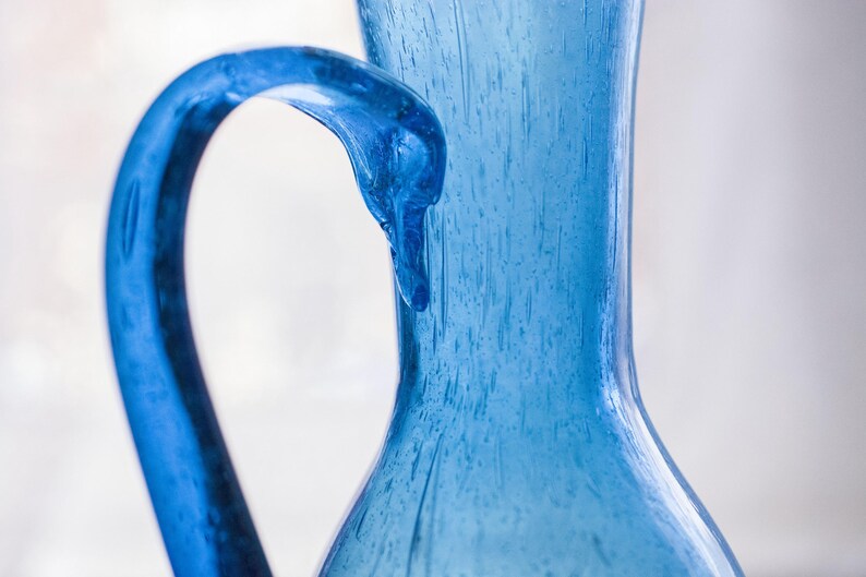 May include: A close-up of a blue glass jug with a curved handle. The jug has a textured surface and a wide opening. The handle is a darker shade of blue than the body of the jug. The background is a soft, blurred white.