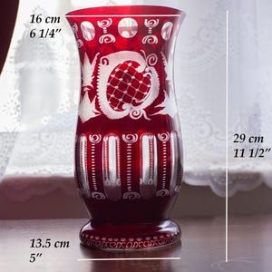 May include: A deep red glass vase, 29 cm tall, featuring white etched patterns. The vase has a flared top, a wide base (13.5 cm), and a detailed rim design. The vase's opening is 16 cm wide.