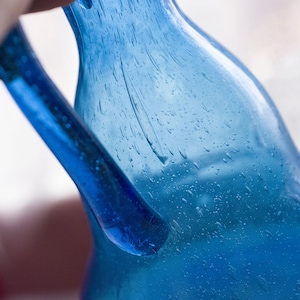 May include: Close-up of a blue glass bottle with a handle. The bottle has a textured surface with small bubbles. The handle is a solid, smooth blue. The bottle is held by a person.