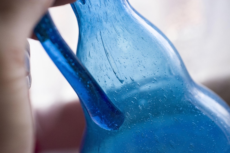 May include: Close-up of a blue glass bottle with a handle. The bottle has a textured surface with small bubbles. The handle is a solid, smooth blue. The bottle is held by a person.