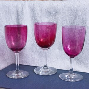 May include: Three magenta-coloured wine glasses with a classic design. Each glass has a rounded bowl and a slender stem. They are arranged on a blue surface against a white background.