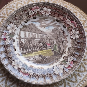 May include: A brown and white ceramic plate with a floral border and a scene of a building with a horse-drawn carriage in the centre. The plate has a scalloped edge and a gold rim.