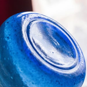 May include: Close-up of a blue glass bottle base. The glass has a textured surface with a glossy finish. The bottle is a vibrant shade of blue, with a circular indentation in the centre.